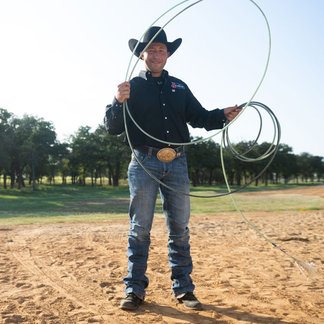 Haven Meged standing in an arena, holding a rope wearing a black shirt and black cowboy hat. 